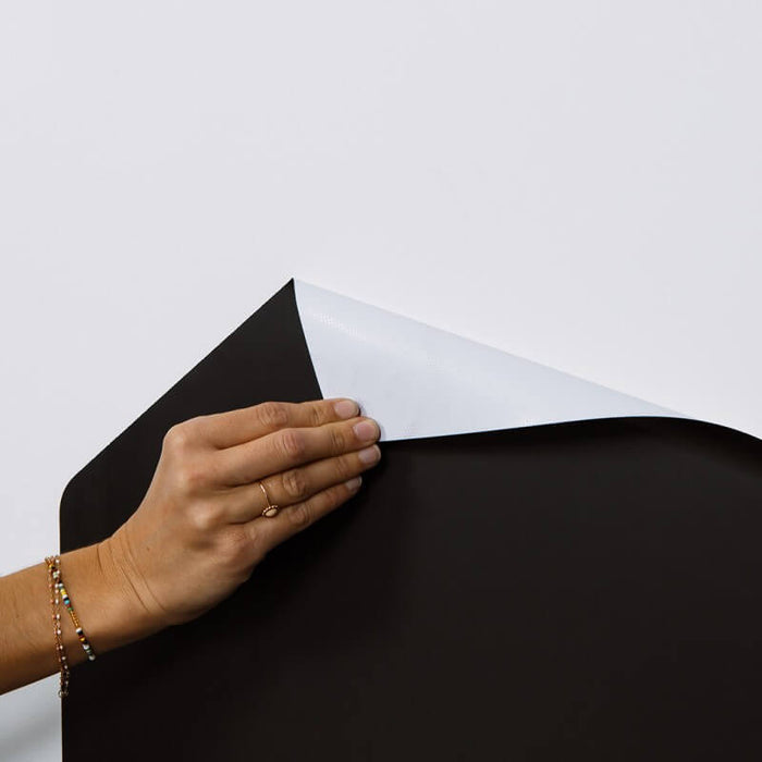 Hand peeling back the corner of a flexible black magnetic chalkboard sheet, showing its white adhesive backing.