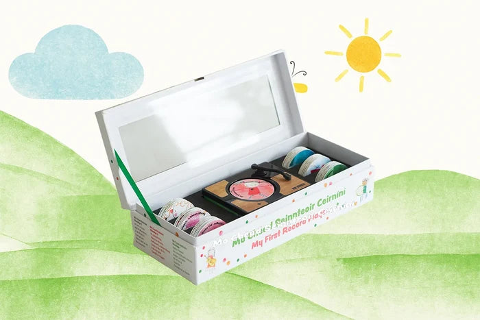 White toy box open to show a wooden record player and colorful discs, set against a sunny landscape illustration.