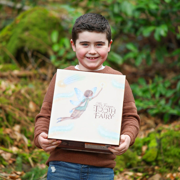 Smiling boy in a brown sweater holds a square gift box with an illustration of a boy tooth fairy with blue wings.
