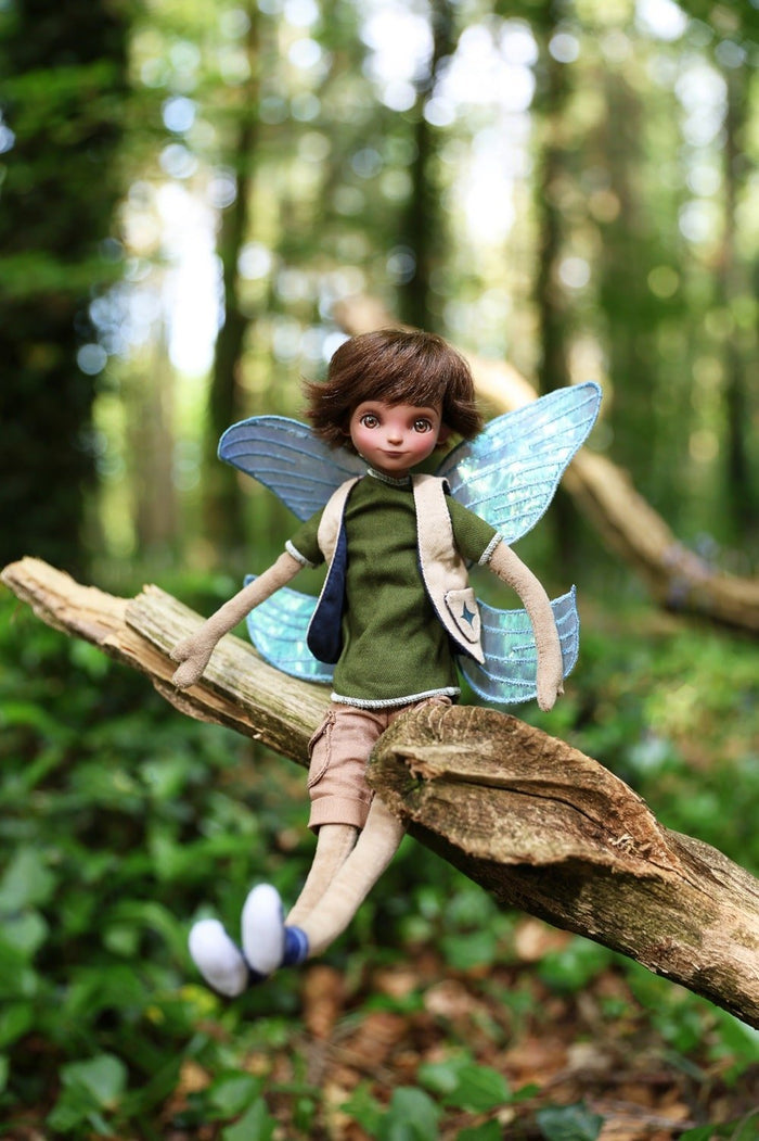Boy fairy doll with brown hair and blue wings sits on a log in a forest, wearing a green shirt and tan vest.