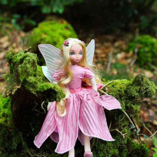 Blonde fairy doll with pink petal-like dress and iridescent wings, sitting on moss-covered rocks in a forest.