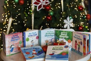Seven colorful Irish board books with cartoon illustrations standing in front of a decorated Christmas tree.