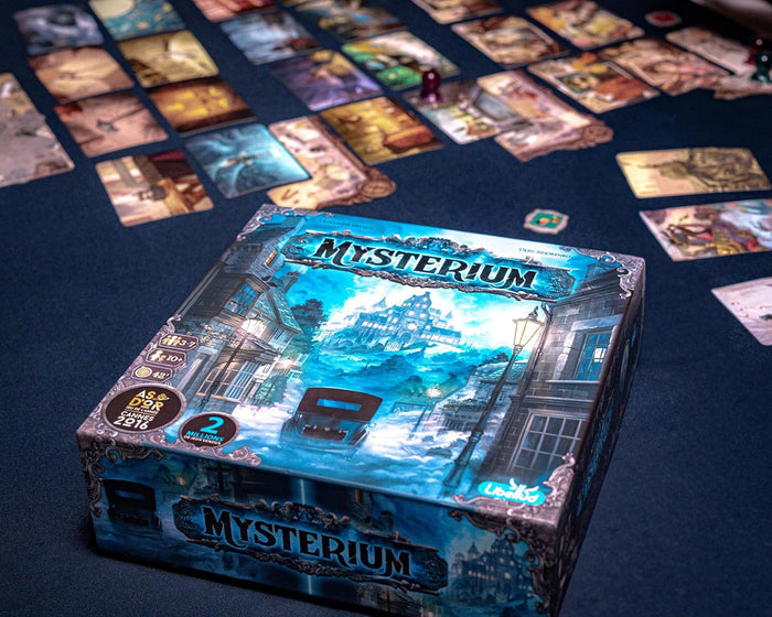 Teal square board game box with mansion and car art, surrounded by spread-out illustrated cards on a dark table.