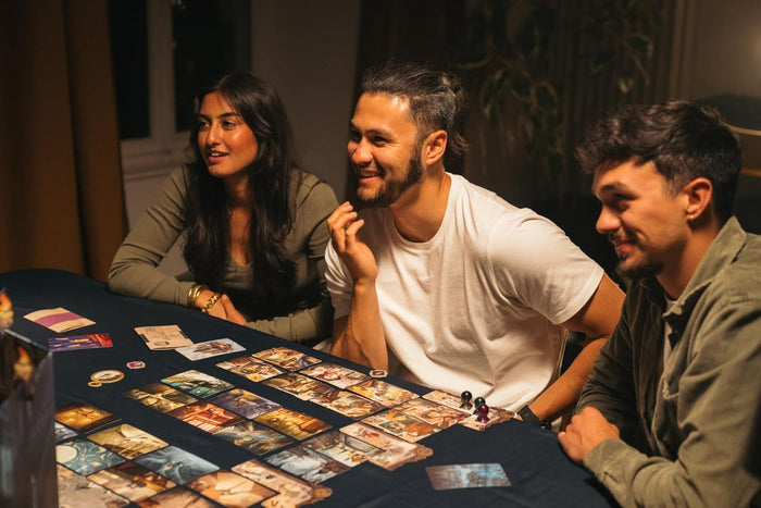 Three people smiling and playing a board game with surreal illustrated cards spread across a dark blue table.