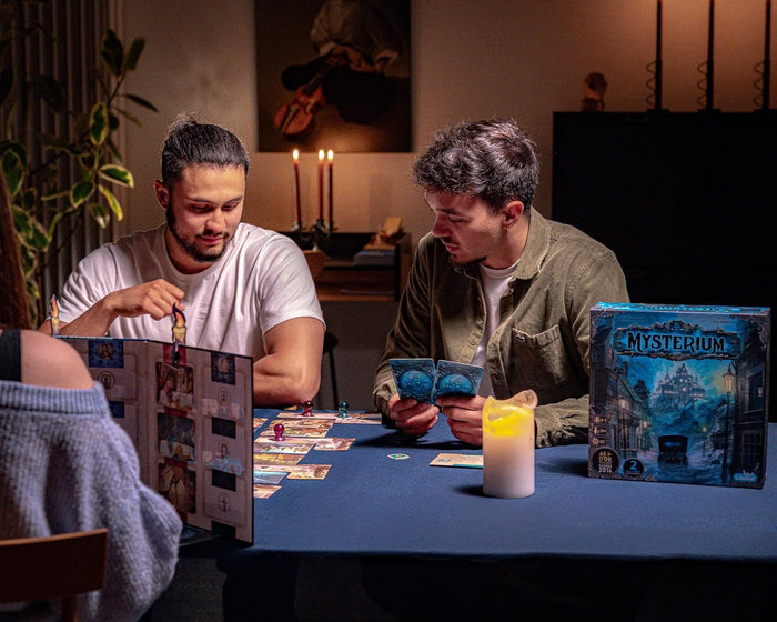 Friends playing Mysterium Refresh board game at a candlelit table with cards, a game screen, and blue box.