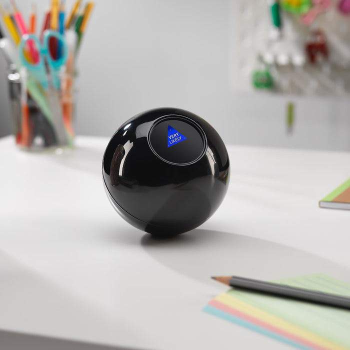 Shiny black sphere showing a blue triangle with VERY LIKELY text, on a white desk with blurred school supplies.