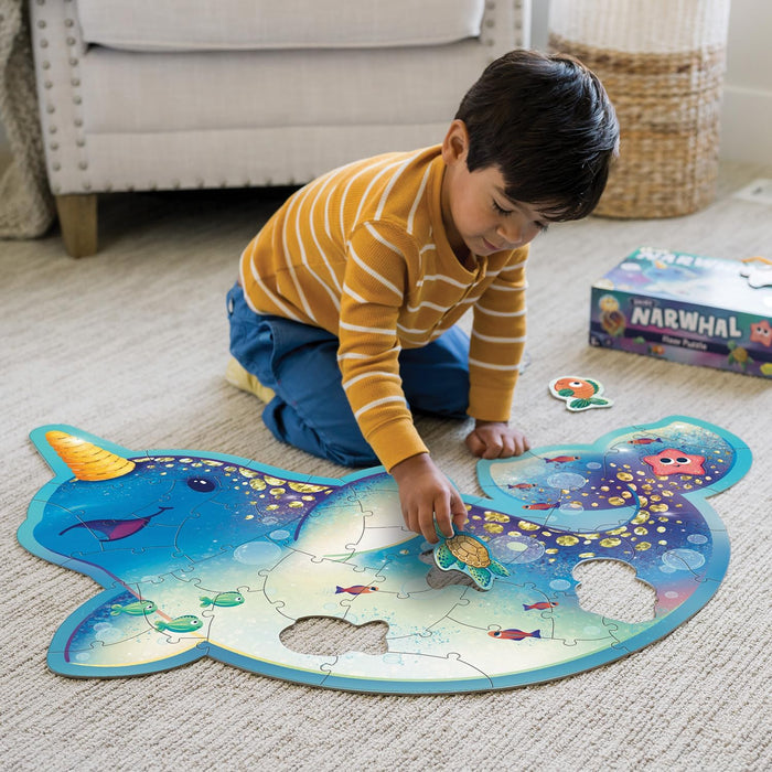 Boy placing a sea turtle piece into a large blue narwhal floor puzzle on a carpet; matching box in background.