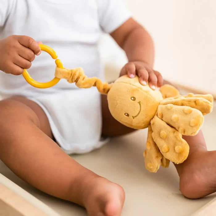 Baby holding an ochre plush octopus toy with textured tentacles and a yellow pull ring.