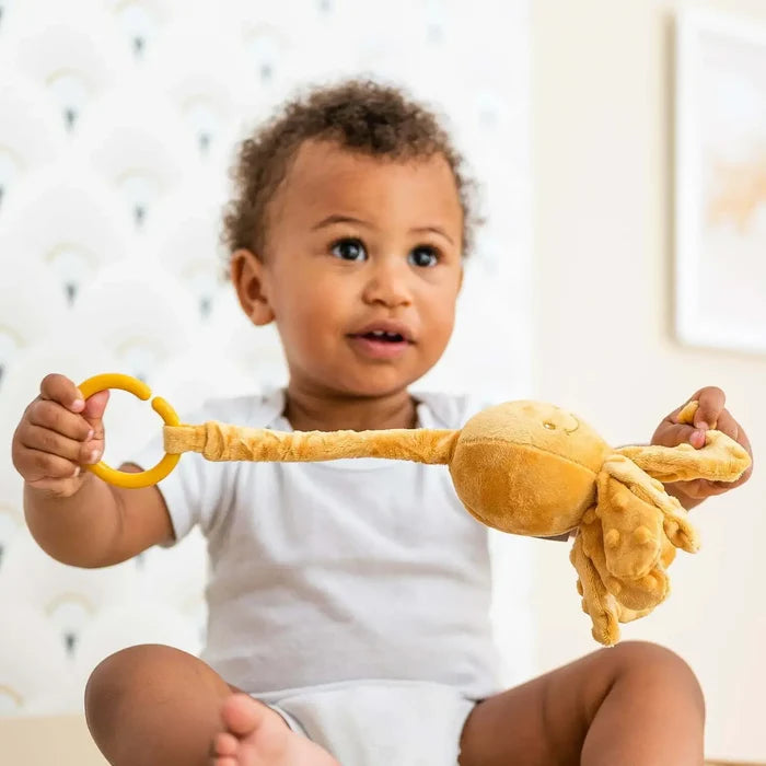 Baby pulls a plush ochre octopus toy with a yellow plastic ring and a long, textured, stretchy neck.