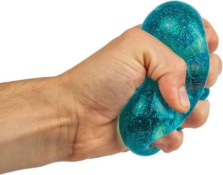 A hand squeezing a translucent teal squishy cube filled with sparkling silver glitter.