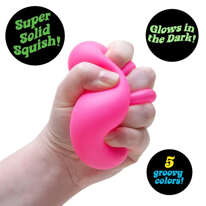 Hand squeezing a bright pink squishy cube. Graphics mention it glows in the dark and comes in 5 groovy colors.