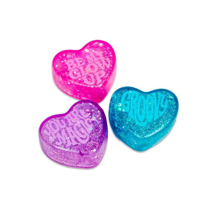 Three glittery heart toys in pink, purple, and blue with 'Be My Glow Up', 'You Are Magic', and 'Groovy' text.