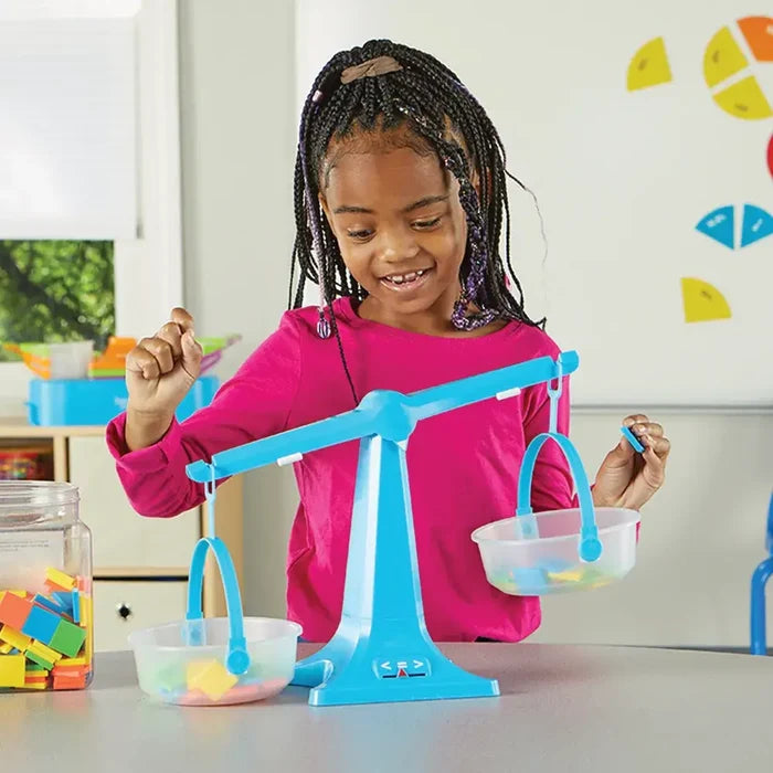 Smiling girl weighing colorful plastic tiles in a blue balance scale with clear hanging pans.
