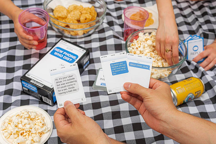 Hands hold text message and blue reply cards from the New Phone, Who Dis? game over snacks on a checkered tablecloth.