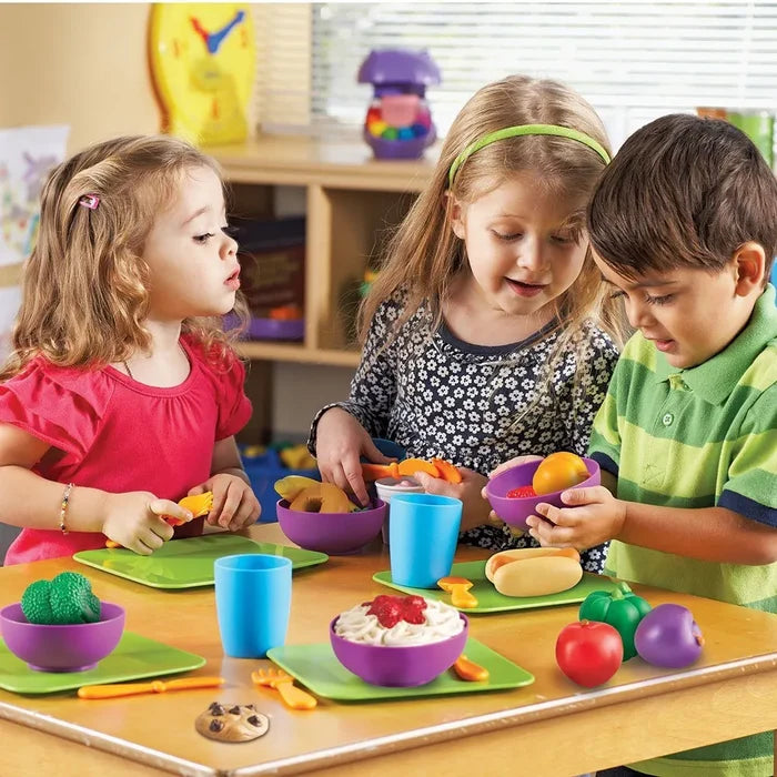 Three kids playing with colorful toy food, purple bowls, green plates, and orange utensils at a small wooden table.