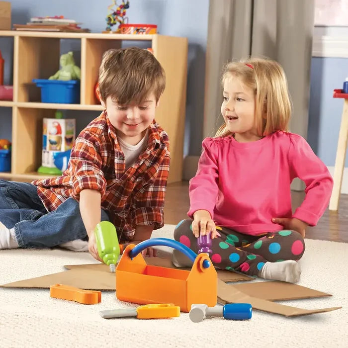 Boy and girl play with a colorful toy tool set featuring an orange toolbox, green drill, and blue hammer on a beige rug.