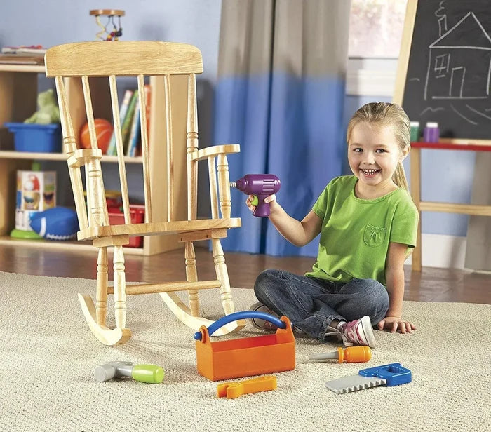 Girl using a purple toy drill on a wooden chair with colorful plastic tools and an orange toolbox on the rug.