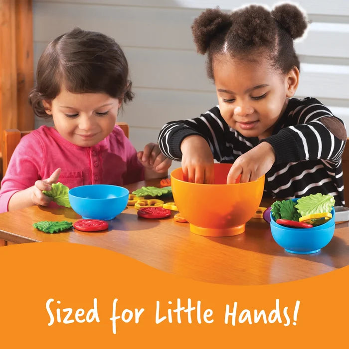 Two children playing with plastic toy lettuce and tomato slices in orange and blue bowls. Text: Sized for Little Hands!