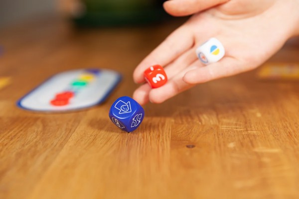 A hand rolls three colorful dice—blue with icons, red with a number 3, and white—onto a wooden table.