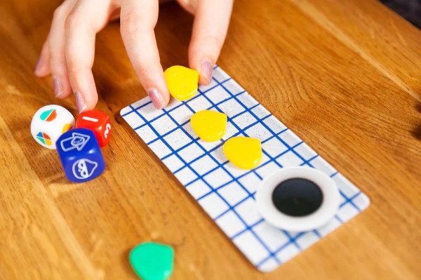 Hand placing yellow bun-shaped pieces on a grid board next to colorful dice and a green token on a wooden table.