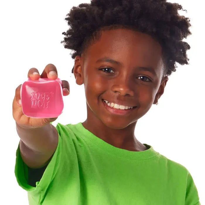 A smiling boy holds a pink, translucent, squishy cube toy with NEE DOH embossed on its surface.