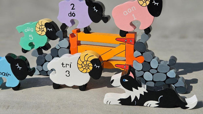 Colorful wooden sheep with Irish number words and numerals, a border collie, orange gate, and grey stone wall pieces.