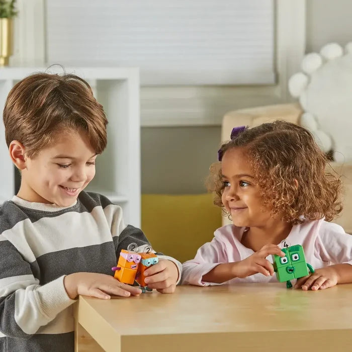 Two children play with two orange block figures wearing glasses and a green square block toy on a wooden table.