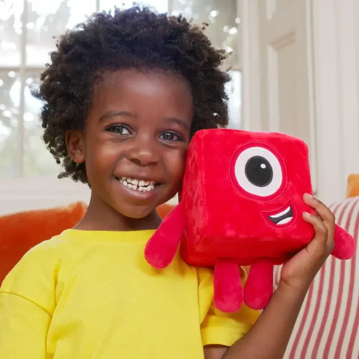 A smiling child holding a soft red cube plush toy featuring one large eye and a friendly, smiling face.