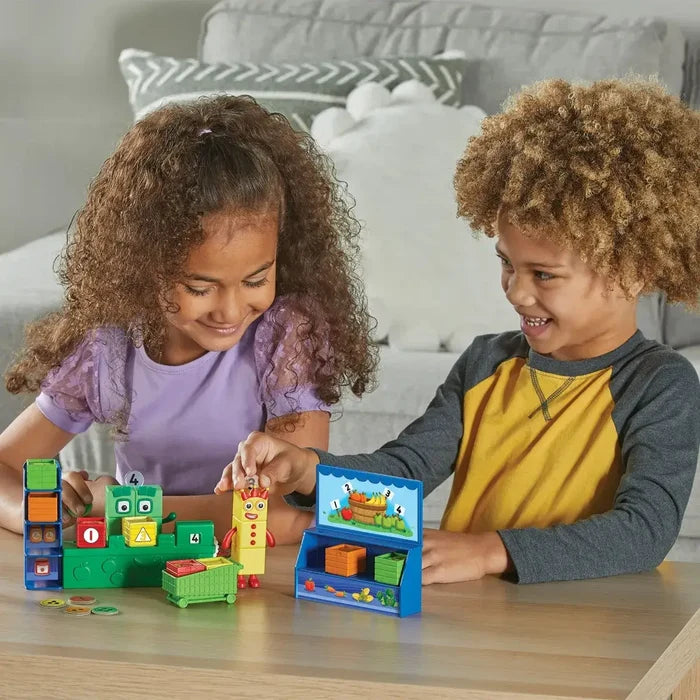 Two children playing with colorful Numberblocks figures, a green toy grocery stand, and a small shopping cart.