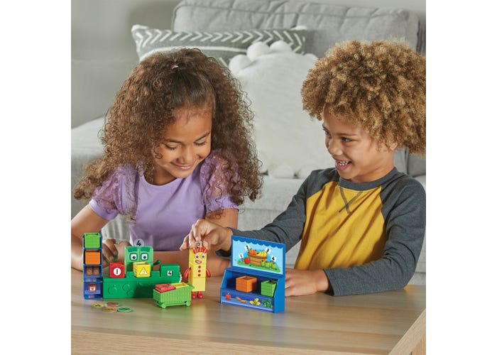 Two children play with a colorful toy market set featuring a blue stall, shopping cart, and numbered block characters.