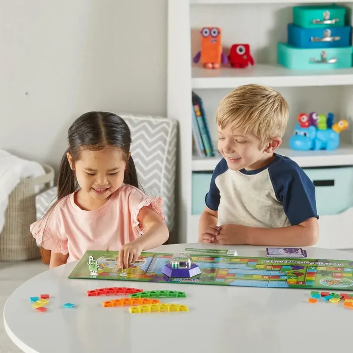 Two children play a colorful board game with pattern bridge pieces and a purple dice popper on a white table.