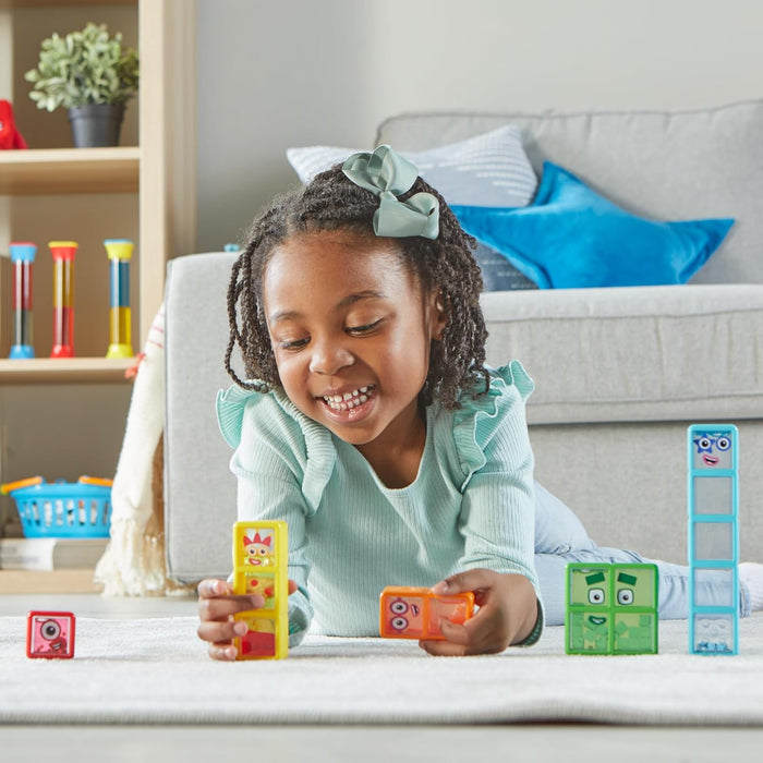 Smiling girl playing with five colorful, rectangular Numberblocks sensory bottles with character faces on a white rug.