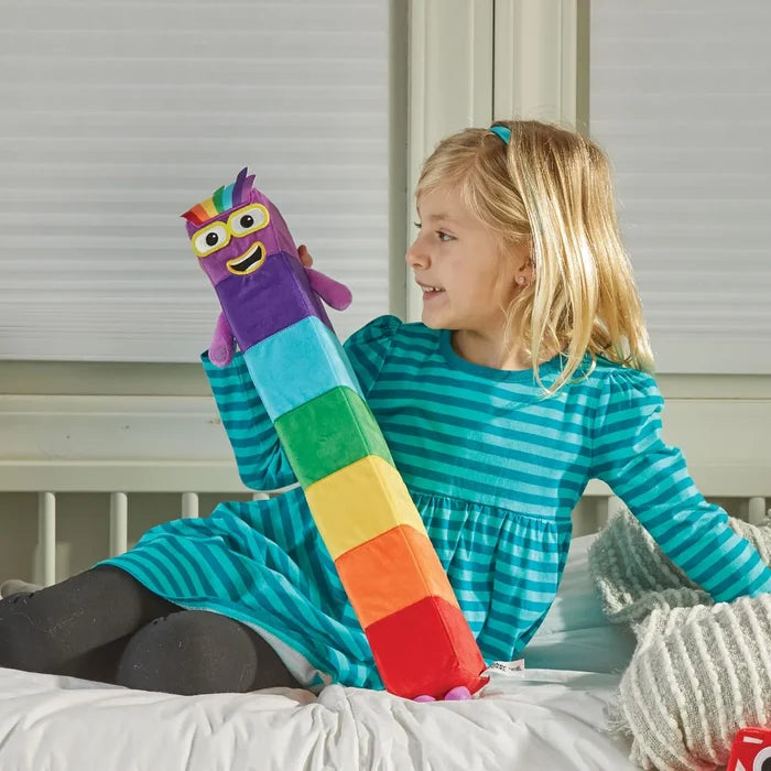 Long rectangular rainbow plush with a smiling face, yellow glasses, and purple limbs, held by a girl on a bed.