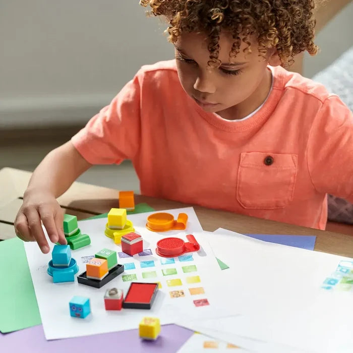 A child using colorful square and round block stamps with matching ink pads to create patterns on white paper.