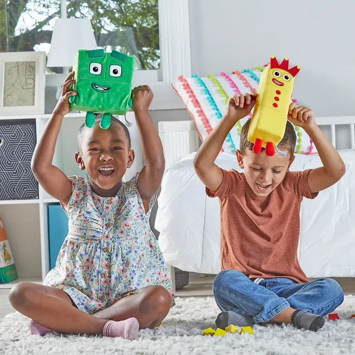 Two kids laugh while holding a soft green square plush and a yellow rectangular plush with a red crown and dots.