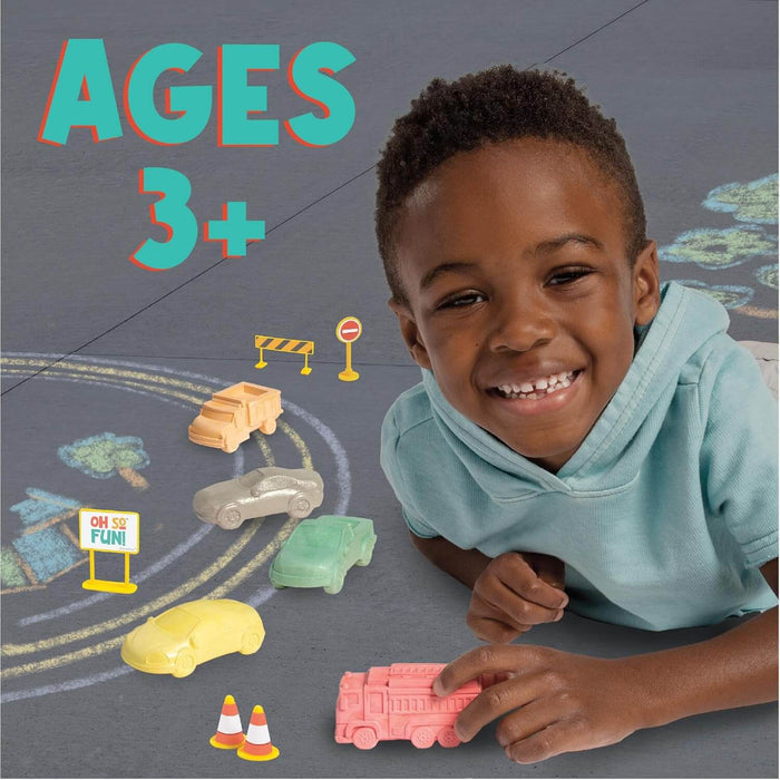 Smiling boy plays with colorful car and truck shaped chalks on a pavement with chalk-drawn roads and toy road signs.