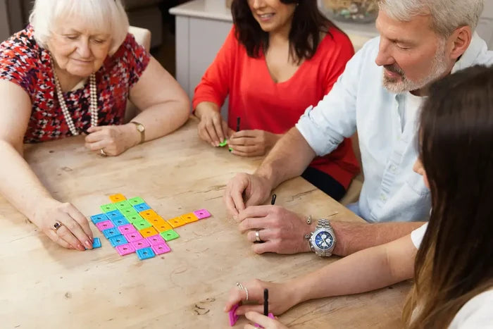 Four people play a tile-placement game with colorful blue, pink, green, and orange square tiles on a wooden table.
