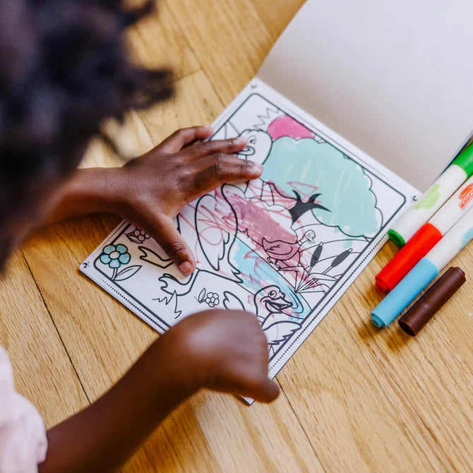 A child colors a duck and pond scene in a farm-themed coloring pad using markers on a light wood floor.