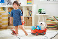 Child pulling a bright orange toy wagon with grey wheels, filled with toys and a stuffed bear, across a playroom rug.
