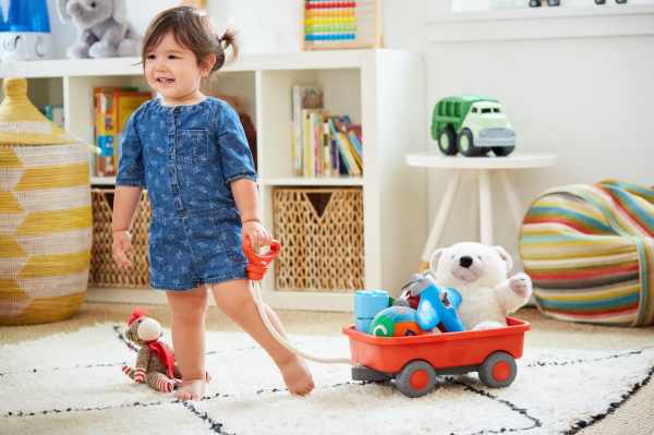 Child pulling a bright orange toy wagon with grey wheels, filled with toys and a stuffed bear, across a playroom rug.