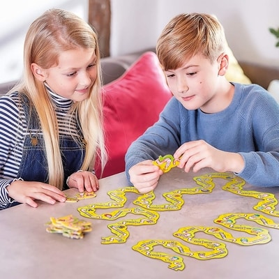 Two children connect yellow, wiggly word tiles to create a long winding path across a tabletop.