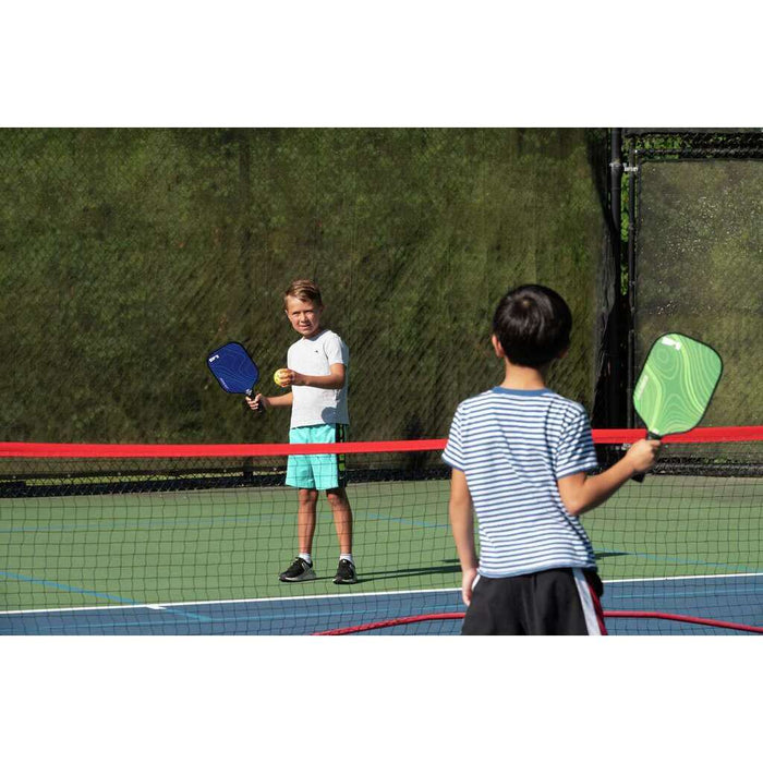 Two kids playing pickleball on an outdoor court with blue and green paddles and a yellow ball.