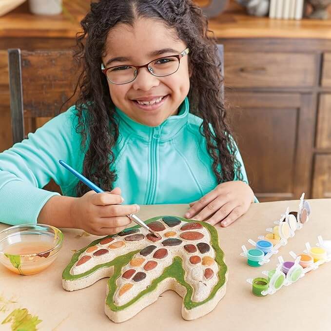 Smiling child painting a dinosaur footprint stepping stone with earth-toned paints and a blue brush on a craft table.