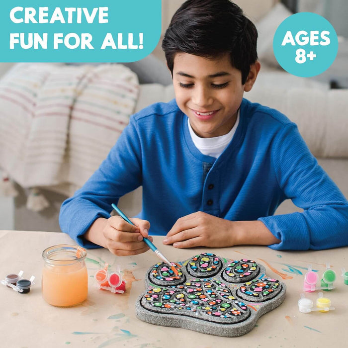 Child painting colorful patterns on a textured grey paw-shaped stepping stone with paint pots and a brush.