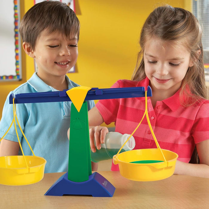 Two children use a green and blue plastic balance scale with two yellow pans to measure green liquid.