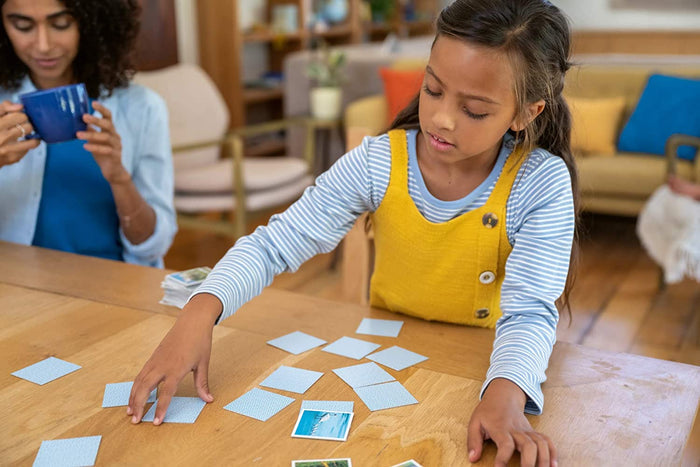 A girl plays a memory matching game with small square cards featuring blue patterned backs on a wooden table.