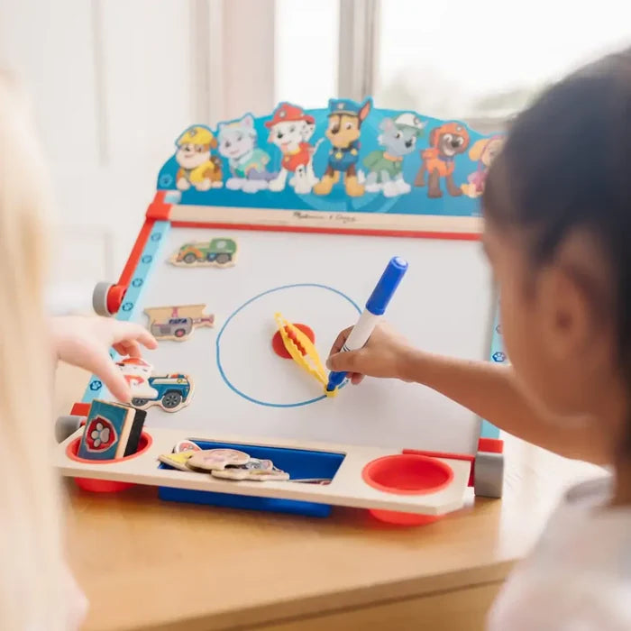 Child draws on a tabletop art easel featuring PAW Patrol character graphics, a whiteboard, and magnetic play pieces.