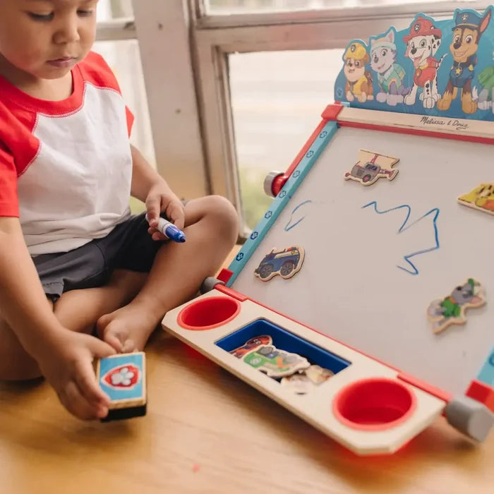 A child plays with a PAW Patrol wooden tabletop easel featuring a whiteboard, magnets, and storage compartments.