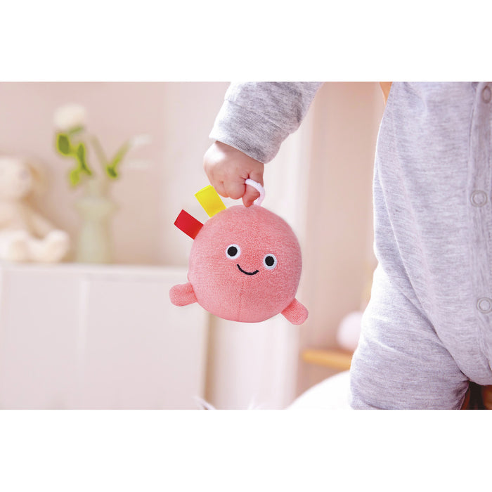 Round pink plush toy with a smiley face and colorful tags held by a baby's hand.