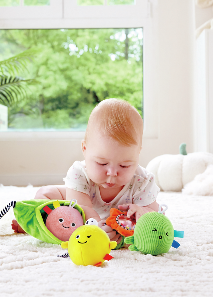 Baby playing with a green plush pod and pink, yellow, and green round characters with cute faces on a white rug.